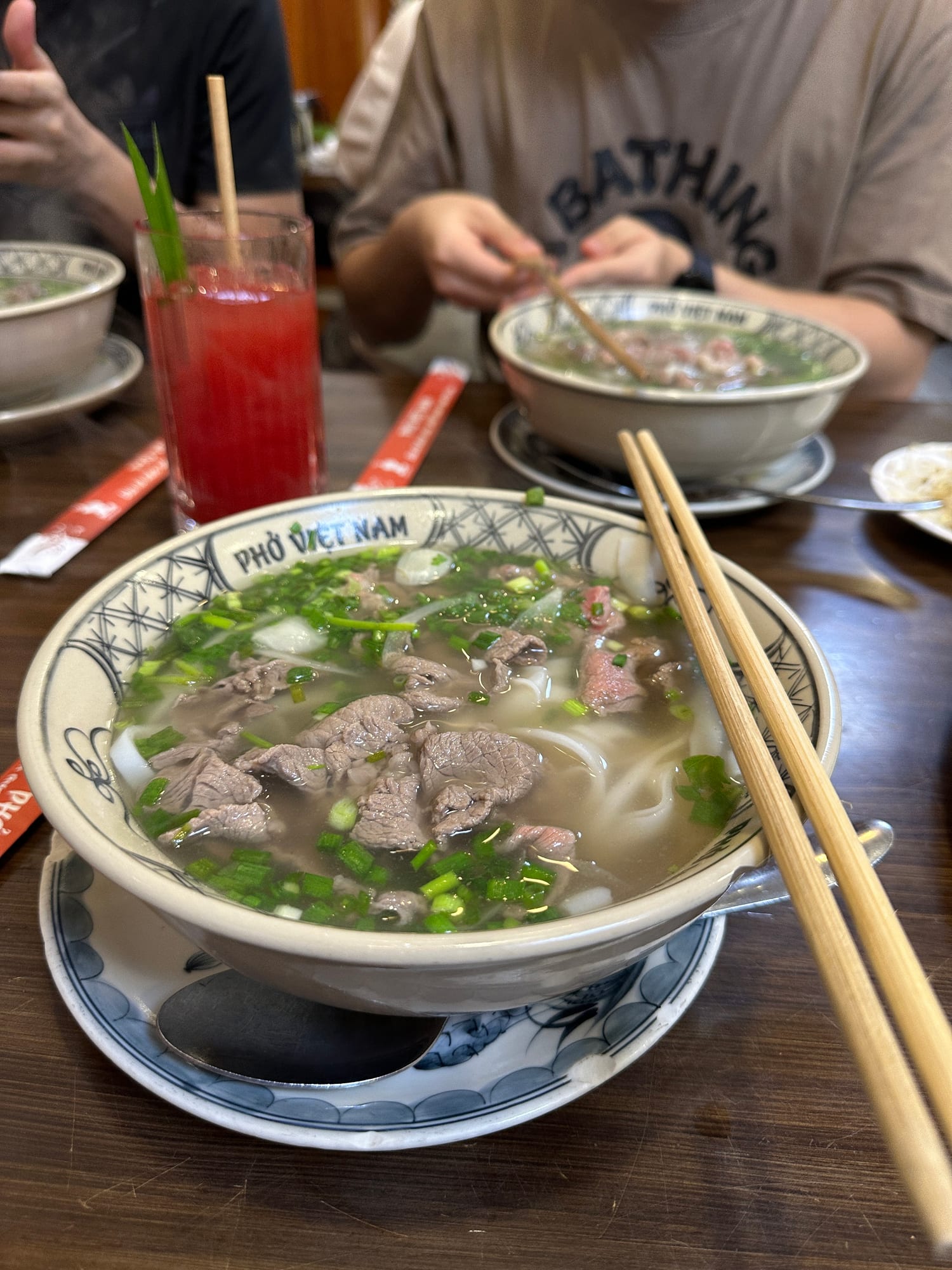 A bowl of phở with beef and rice noodles at a Phở Phố table with diners in the background